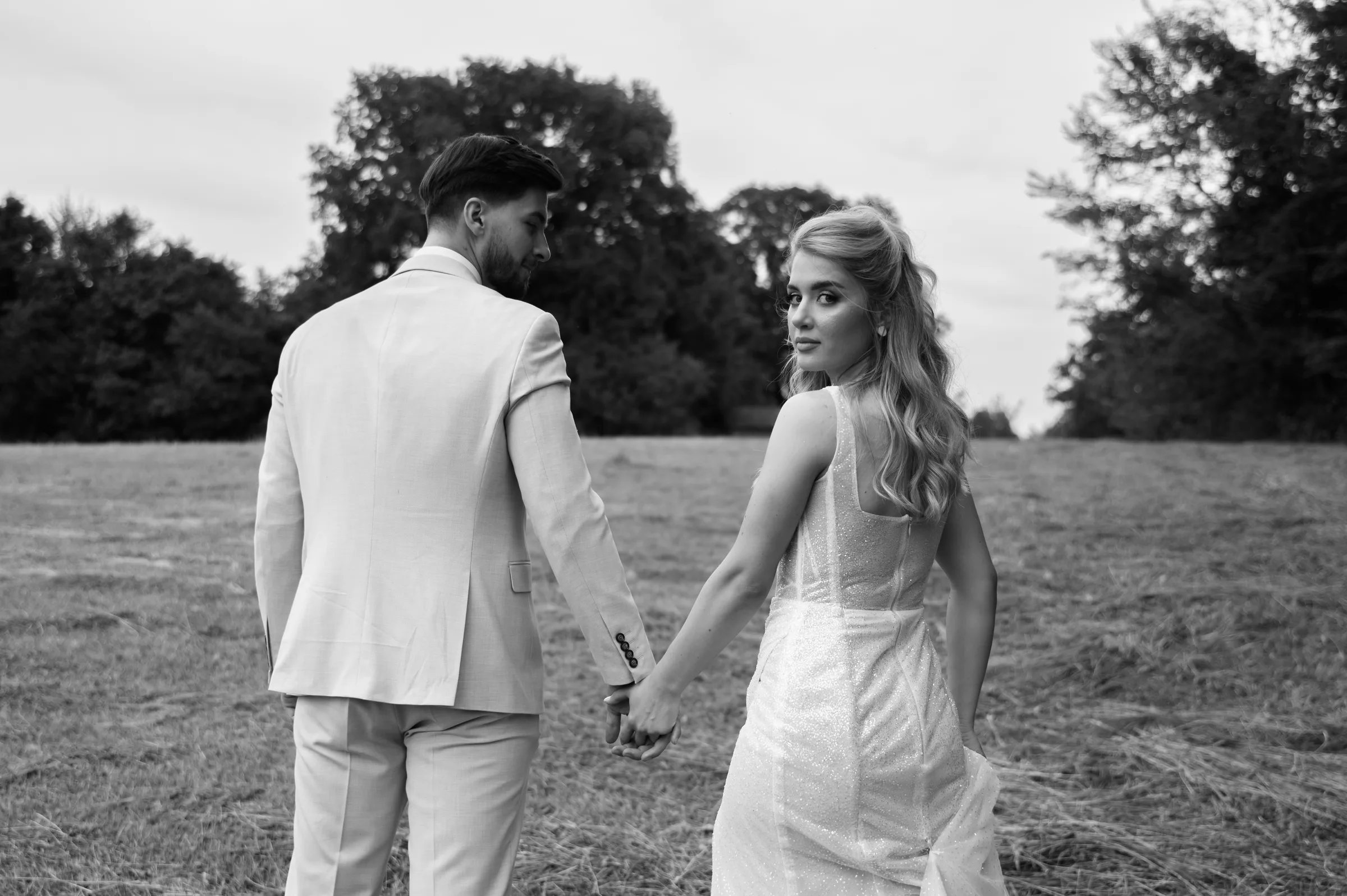 Mladenci se drže za ruke i šetaju kroz polje — MI Weddings.